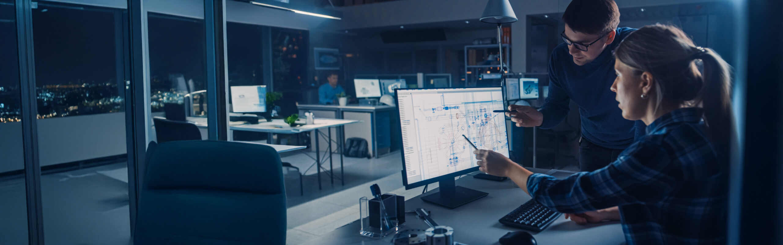 Kaksi ihmistä toimistossa, jotka keskittyvät tietokoneen näyttöön, jossa näytetään kaavioita tai tietoja. Toimisto on moderni, ja sen suurista ikkunoista näkyy öinen kaupunkikuva, ja taustalla työskentelee muita ihmisiä.
